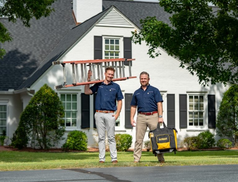 Two Parahome Solutions technicians carrying a ladder and toolbox in front of a white two-story home
